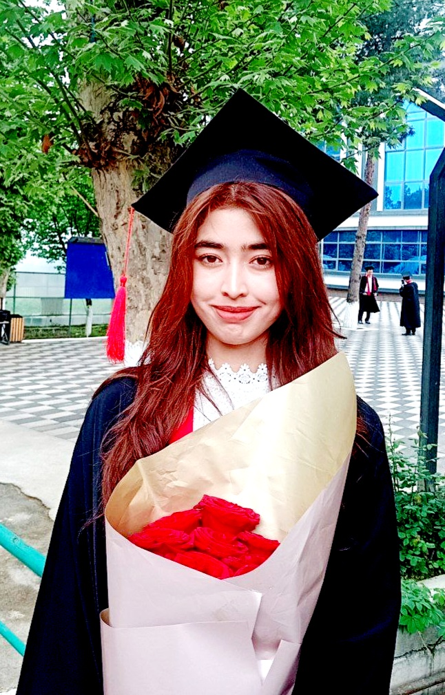Young Central Asian woman in a graduation cap and gown holding a bouquet of flowers. She's in front of a tree and a large school building.