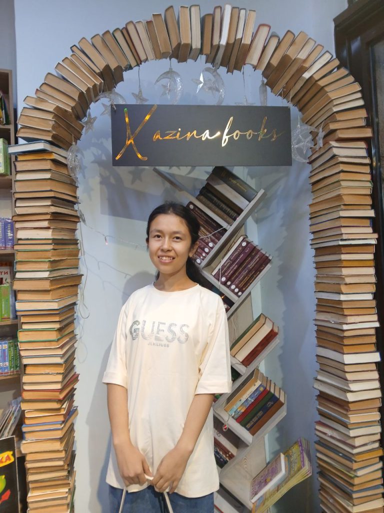 Central Asian teen girl in a white tee shirt, long black hair in a ponytail, an arch of books behind her. 