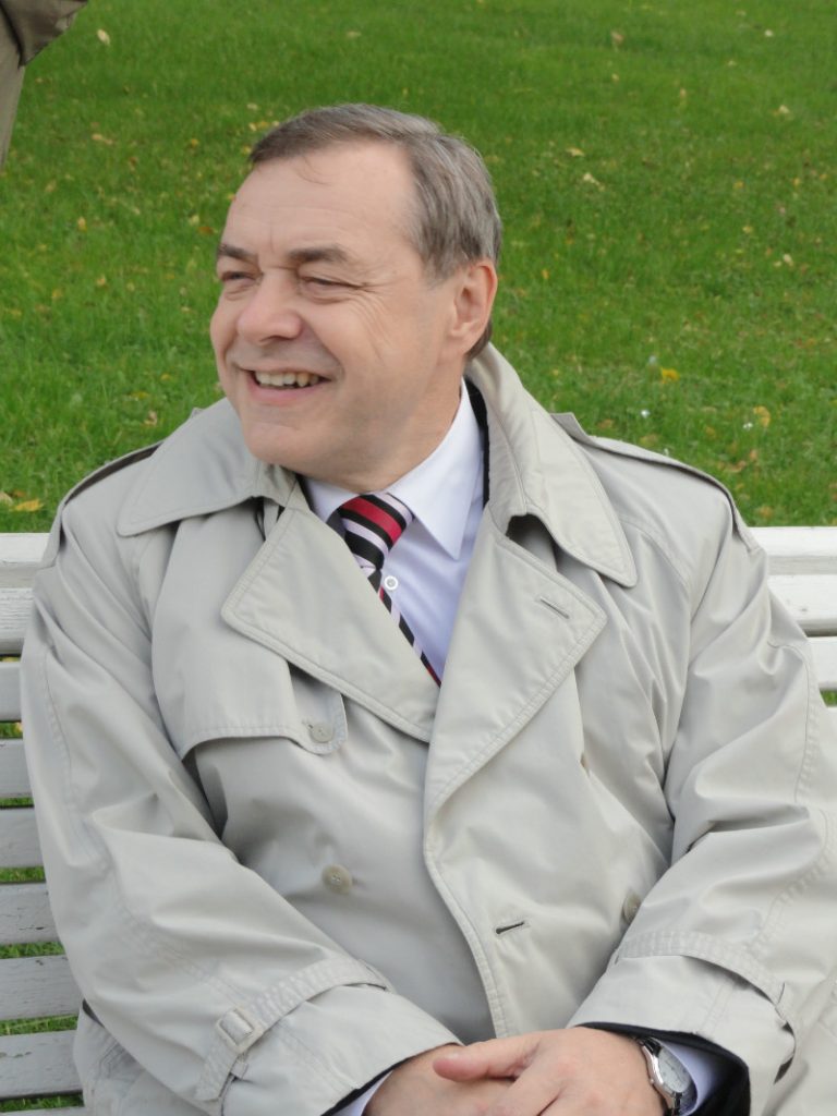 Older white man with thinning gray hair, a gray jacket, and a white shirt and tie on a bench with a green lawn in the background. 