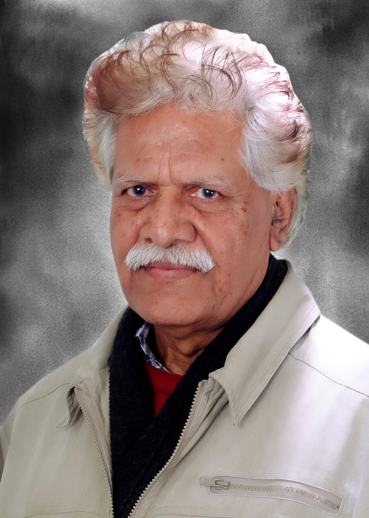 Older South Asian man with white curly hair, a neatly trimmed mustache, and a brown zipped coat and collared shirt and red sweater.