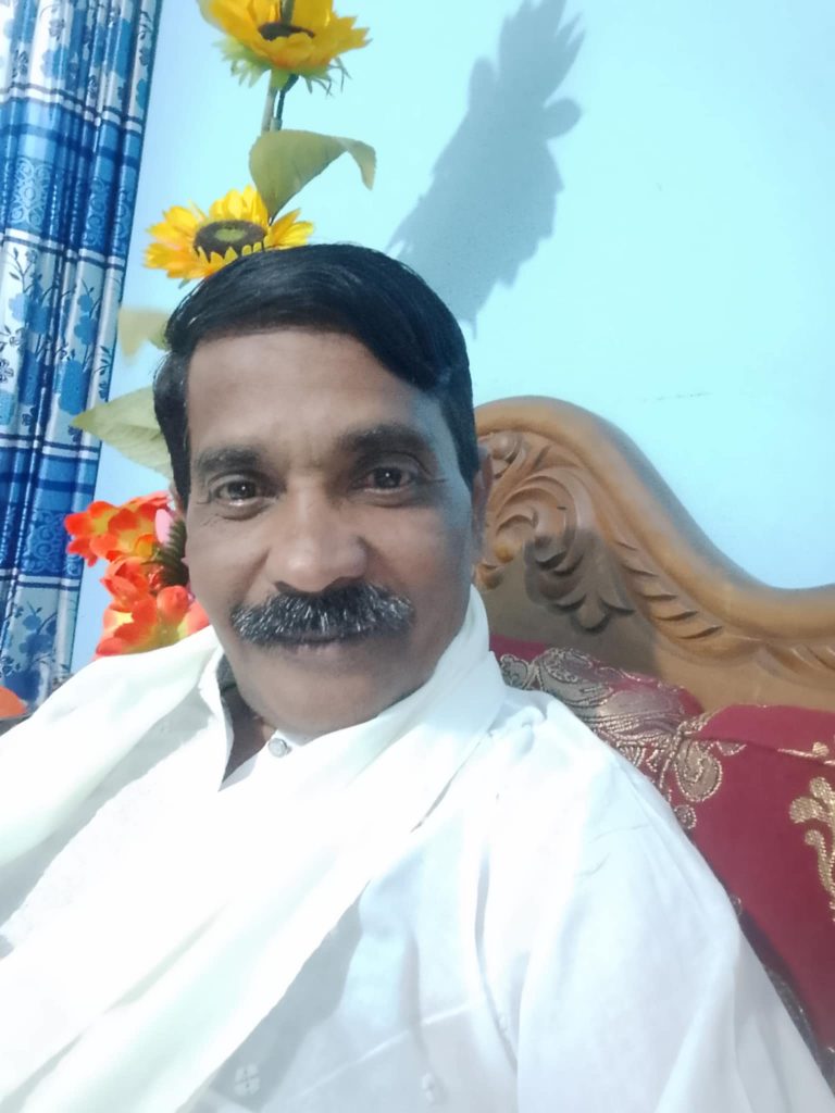 Middle-aged South Asian man with a mustache, no beard, and a white collared shirt sitting in a wooden chair with a pillow. Flowers and curtains are behind him.