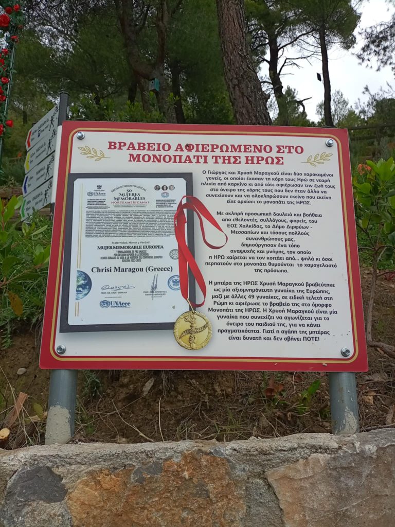 Sign in Greece in red and black and white marking the entrance to a hiking trail known as "The Hero's Path." Trees and bushes in the background, cloudy day. 