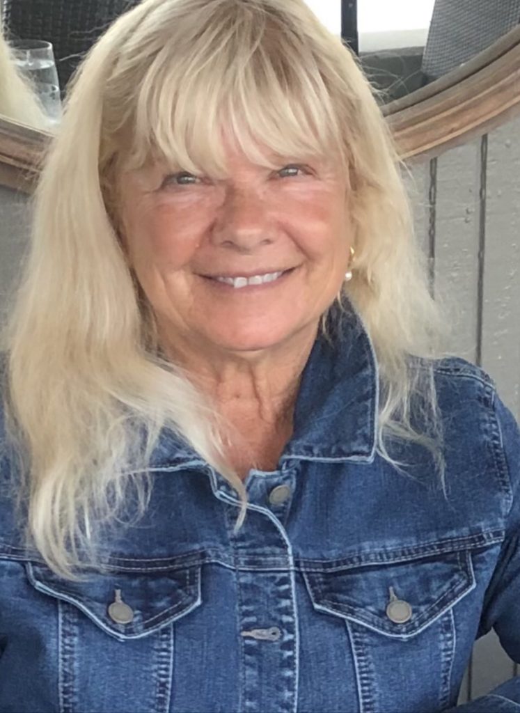 Middle aged white woman with blonde hair, green eyes, earrings, and a blue denim jean vest.