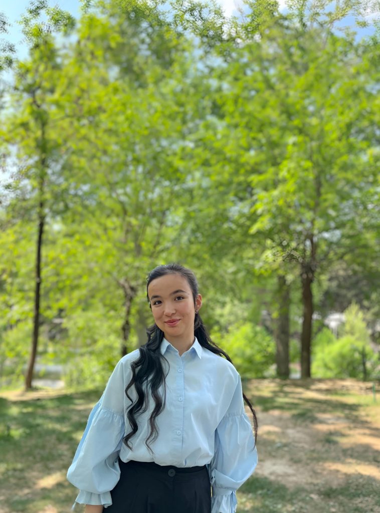Central Asian teen girl in a light blue collared ruffled blouse and black skirt in a grassy field with leafy green trees.