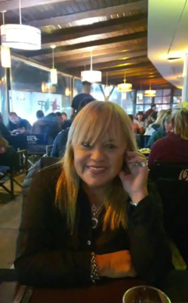 Middle-aged Latina woman with straight light brown hair, brown eyes, a few rings on her fingers, a necklace, bracelet, black jacket, and a smile. She's seated at a restaurant table. 