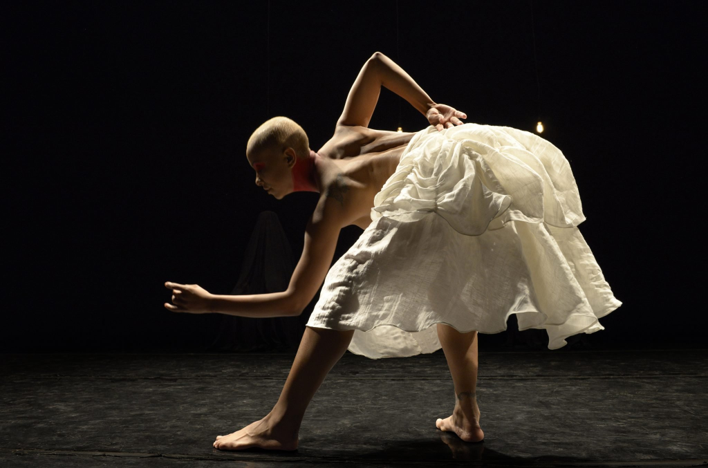 Bald person in a white ruffled tutu bending over to the left in a profile view.
