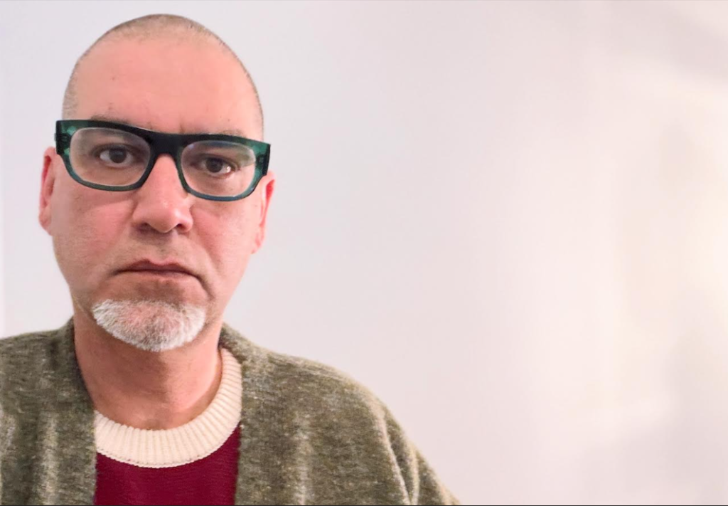 Middle-aged bald white man with a small white beard, reading glasses, a red and white sweater, and gray sweater over that.