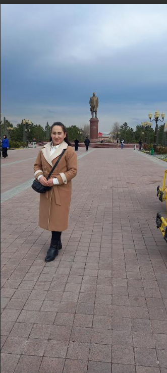 Young Central Asian woman stands on a concrete path with trees and a statue in the background. It's a cloudy day.