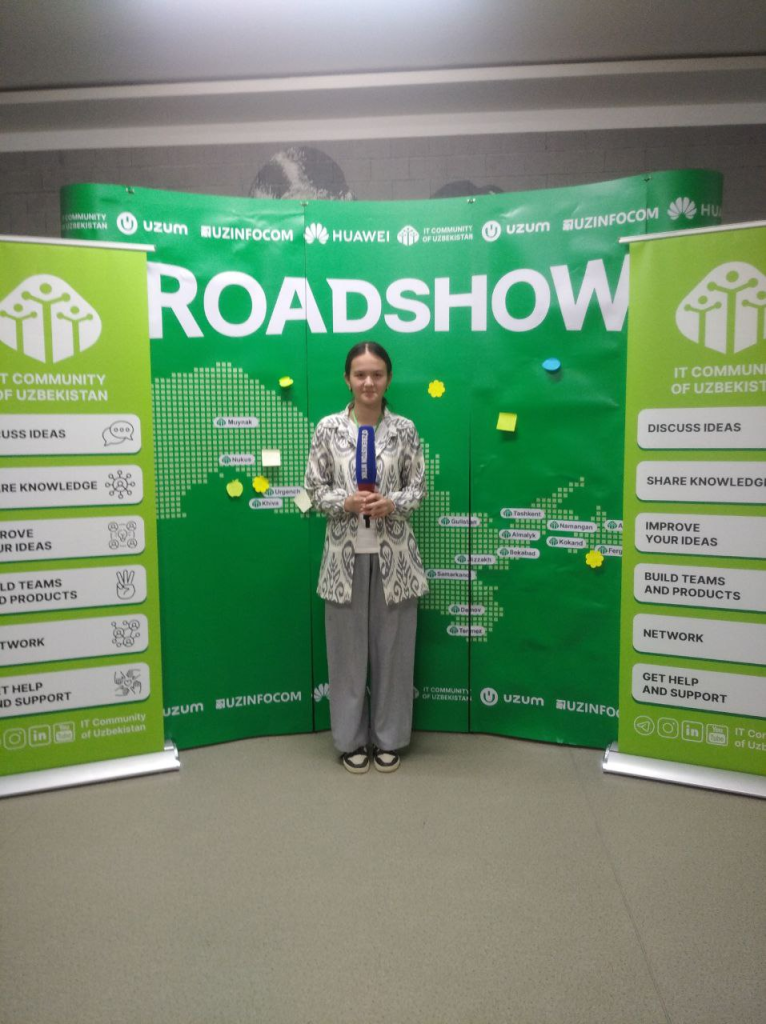 Young Central Asian woman stands in front of a tech billboard at a product expo. She's in a paisley jacket, long dark hair up behind her head, and gray slacks. 