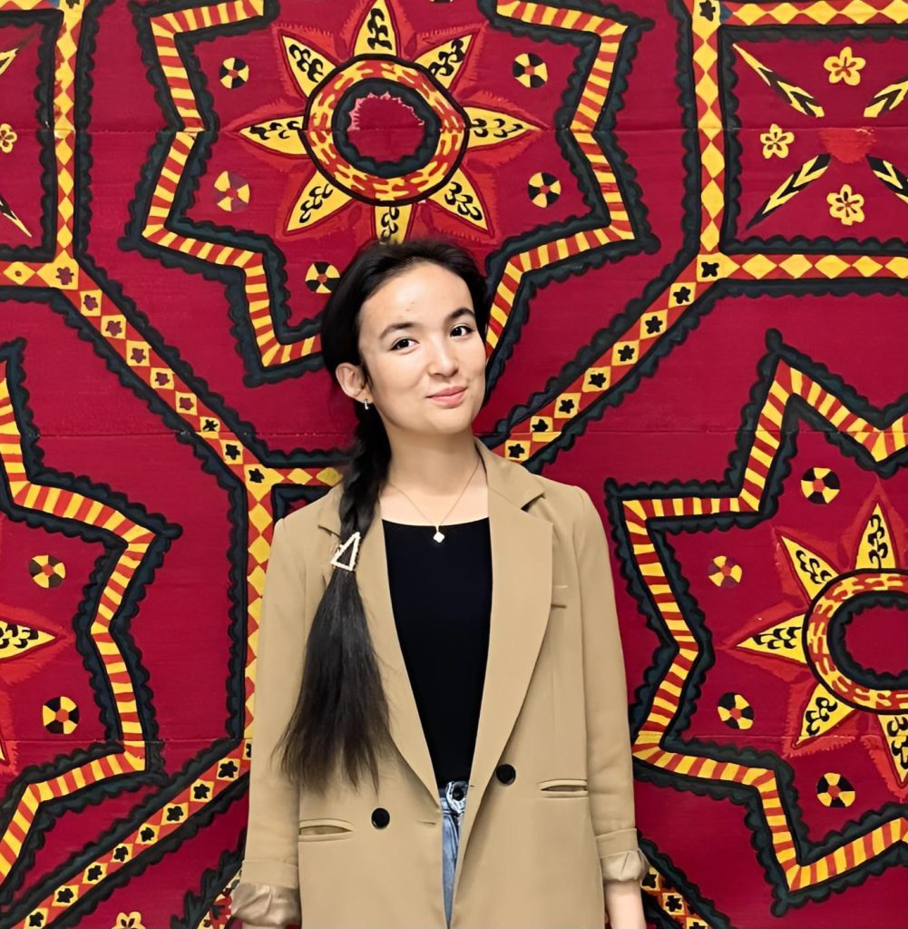 Young Central Asian woman with a long dark braid, black top and brown coat. Bright red quilt with yellow and black patterns behind her. 