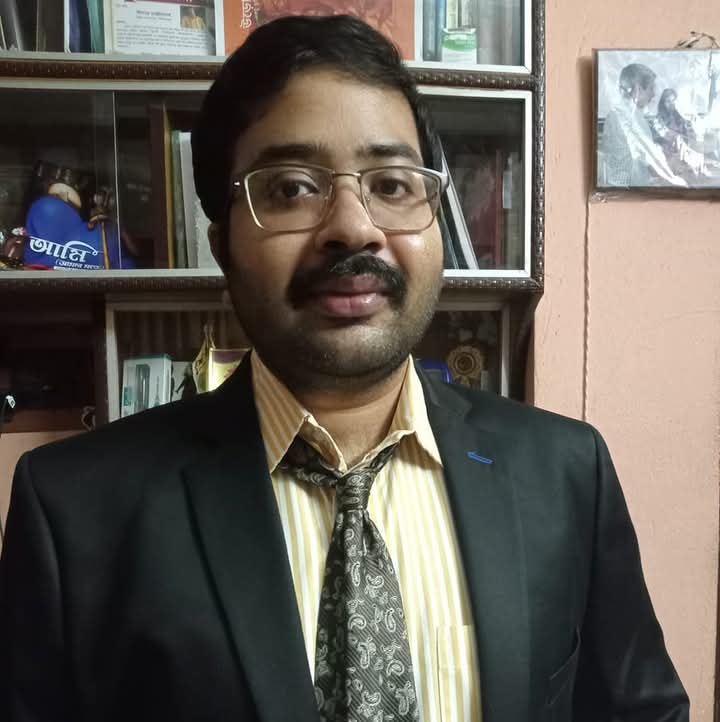 South Asian man, young middle age, clean cut, reading glasses, black coat, yellow and white striped shirt, patterned purple tie, standing in front of a bookshelf.