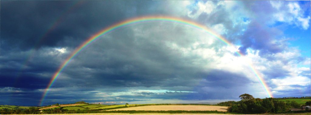 Double rainbow in a blue cloudy sky over the mostly flat English countryside, plains and trees.