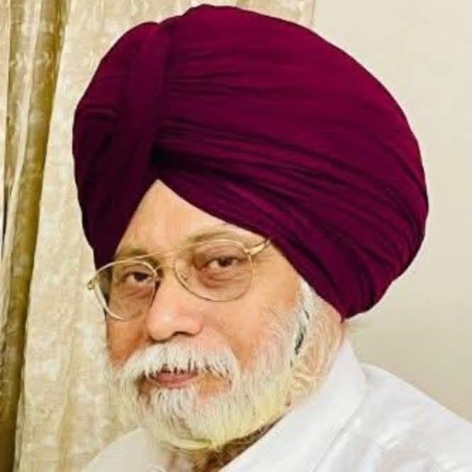 Older South Asian man with reading glasses and a purple turban and beard and white shirt.