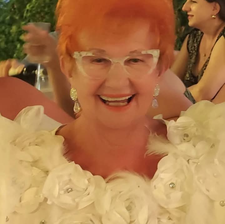 European woman in a fluffy floral white dress, double pendant earrings, reading glasses, and red hair.