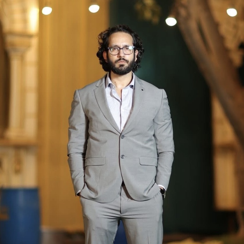 Middle-aged South Asian man in a gray coat and white collared shirt with dark curly hair and reading glasses standing in front of a large concrete building at night.