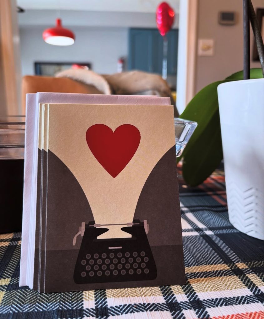 Indoor table with a houseplant with green leaves, a doorway and lamps, and stationery with a red heart. 