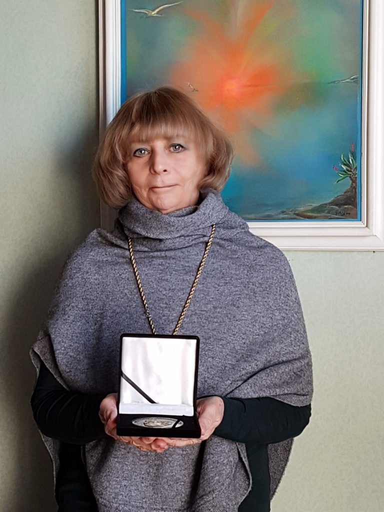 Middle aged European woman with light brown hair, a long necklace, and a gray sweater standing in front of a red and blue painting of water and a beach. She's holding an award. 