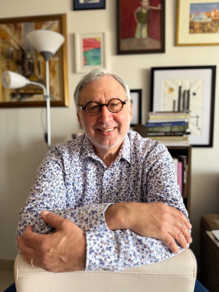 Older light skinned man with reading glasses and a white collared shirt with a blue floral pattern seated at a desk. Books, abstract paintings, and a lamp are behind him. 