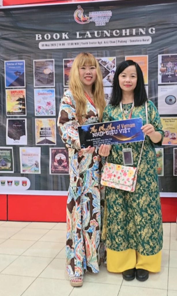 Author Vo Thi Nhu Mai, right, in a green dress with yellow flowers and a pink and white and yellow purse standing next to another woman in a dress and patterned pink, brown, and blue dress in front of a display of book colors.