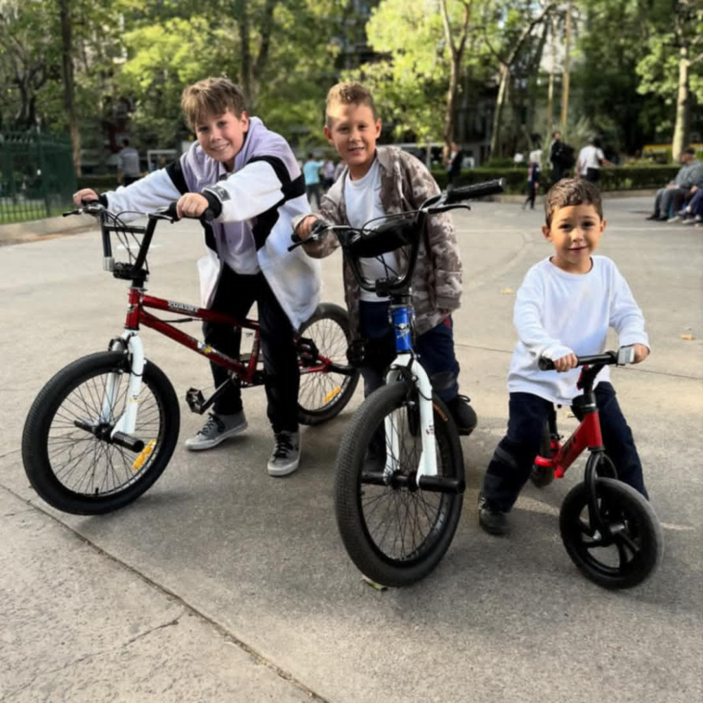 Three light skinned boys in white shirts and jackets and black pants on children's bikes in a park area with nearby green trees.