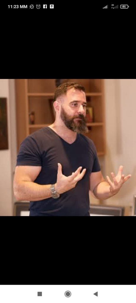 European man, light skinned with a trimmed brown beard, short hair, brown eyes, and a black tee shirt and watch. Hands out in front of him as if he's explaining something. 