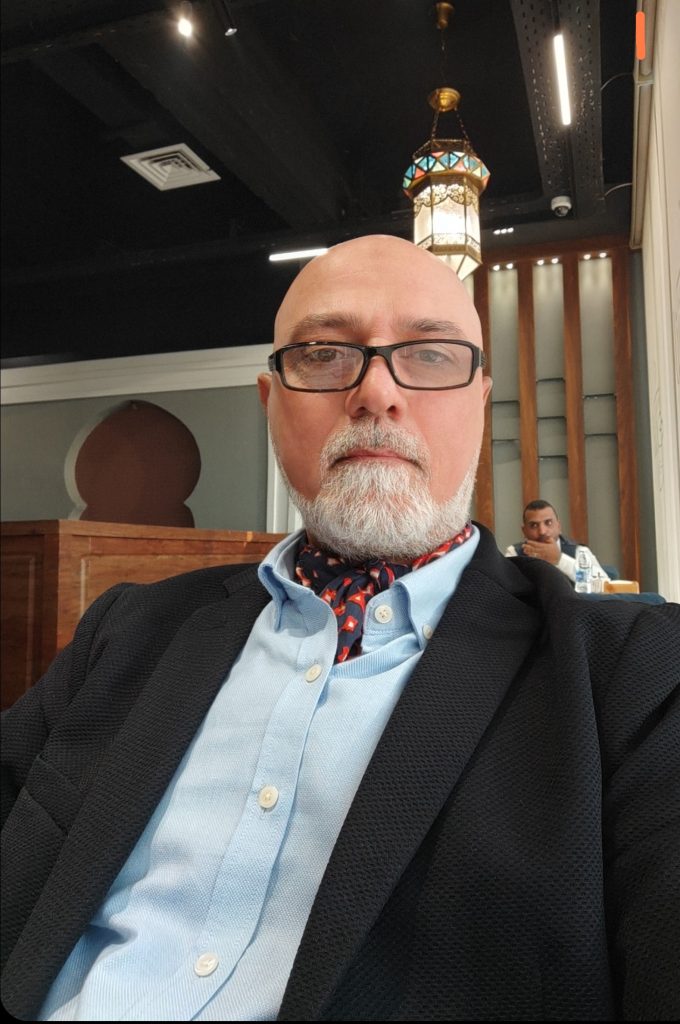 Older bald man with reading glasses, a white trimmed beard, a blue shirt and a grey coat and a red and black tie.