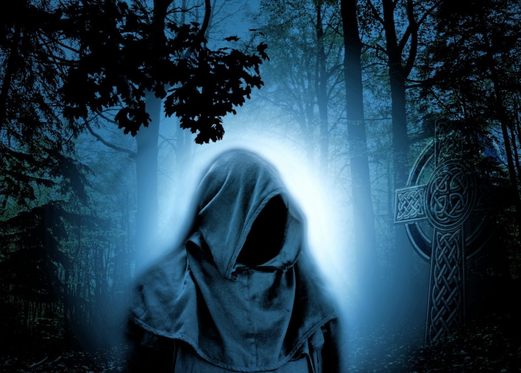 Hooded bodiless figure in a graveyard at night, lit up by moonlight. Trees and foliage and a giant Celtic cross tombstone.