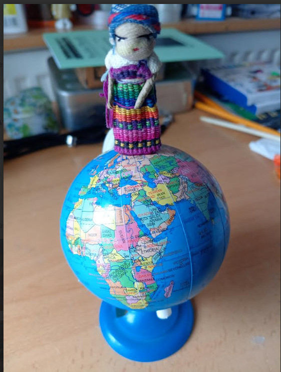 Woven doll figure on top of a globe, resting on the North Pole. Globe is on a desk with other writing and art implements.