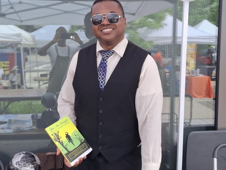 Young middle aged Black man in a white collared shirt and blue patterned tie, sunglasses and a black vest, holding a copy of his book You Are Enough at an open air book fair.