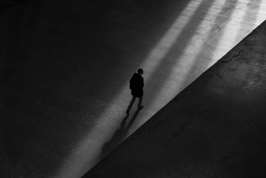 Solitary figure with skinny legs and a backpack navigates an empty room towards a beam of light. Black and white image aerial view.