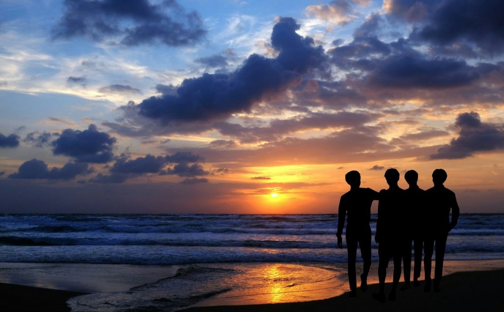 Group of silhouetted people on a beach at sunset or sunrise. Yellow sky behind the clouds near the horizon, water heading out at low tide.