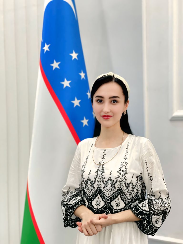 Young Central Asian woman with a white headdress, long dark hair, brown eyes, and a white and black patterned top standing in front of the Uzbek flag.