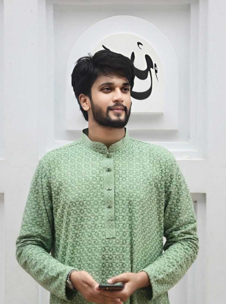 Young South Asian man with a trimmed beard and mustache, short dark hair and a light green collared shirt. He's standing in front of a white wall with an Arabic character behind him.