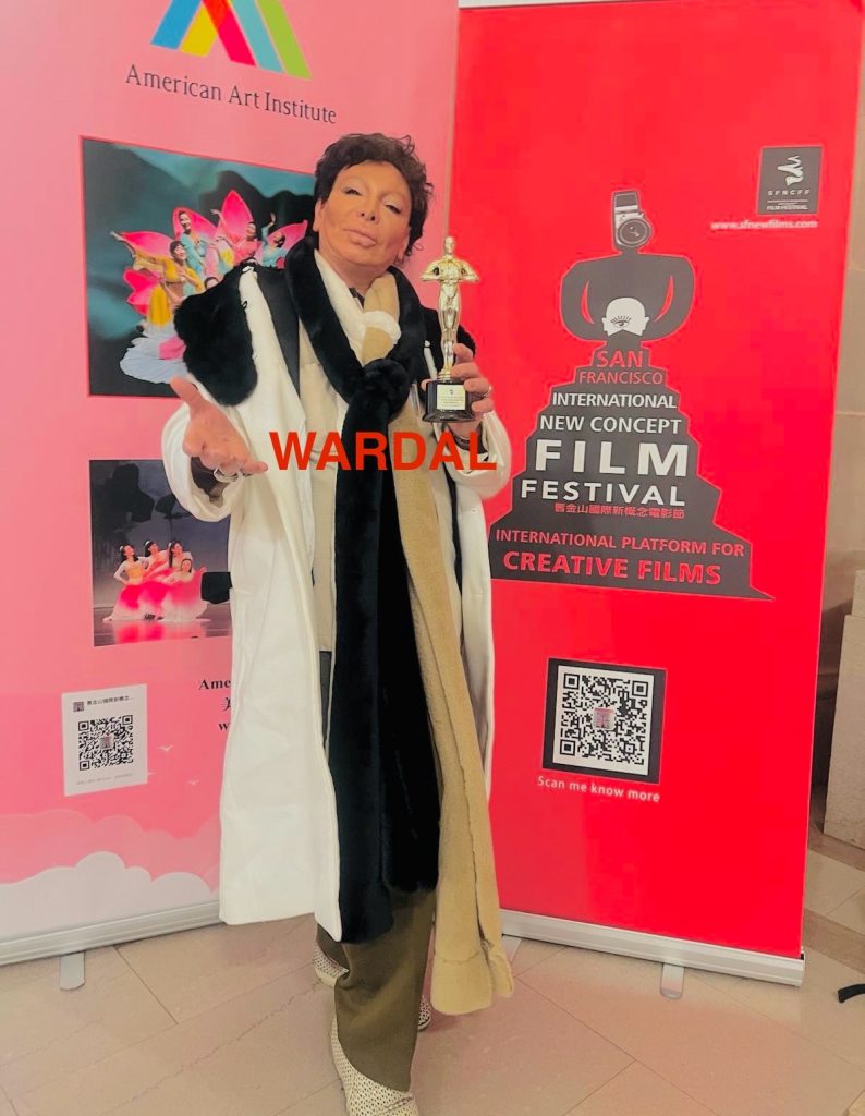 Artist Federico Wardal, a middle aged brown haired Italian man, in a white coat and black scarf, holding a film trophy from the San Francisco International New Concept Film Festival. He's in the corner of pink and red walls at the American Art Institute. 
