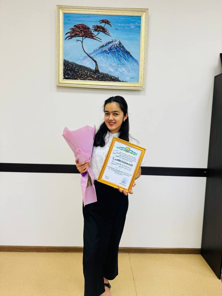 Young Central Asian woman with dark hair, a white blouse and black skirt holding an award and bouquet of flowers with a painting of trees and mountains above her. 