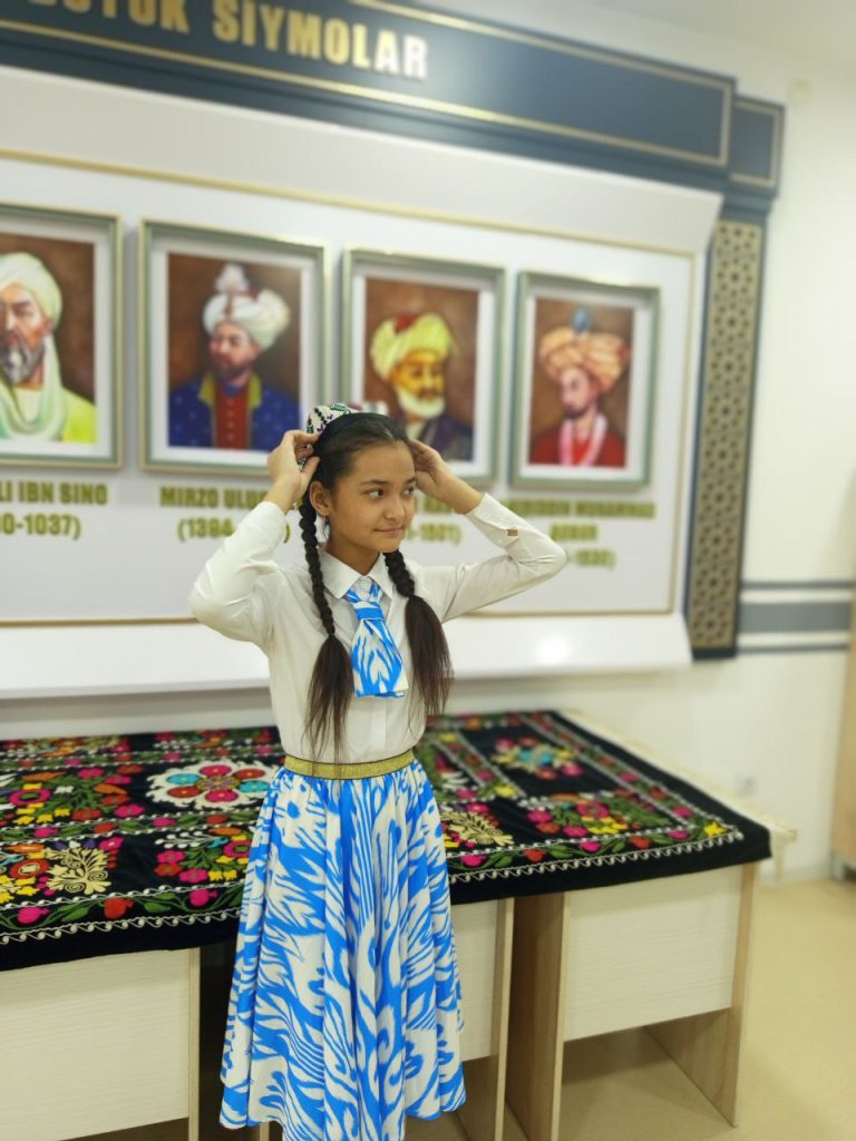 Young teen Central Asian girl with an embroidered headscarf and blue and white scarf with long dark hair in front of a photo gallery with images of historical Uzbek leaders. 