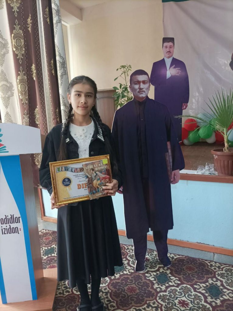 Young Central Asian teen girl posing with an award and a book in a room with cutouts of her national leaders.