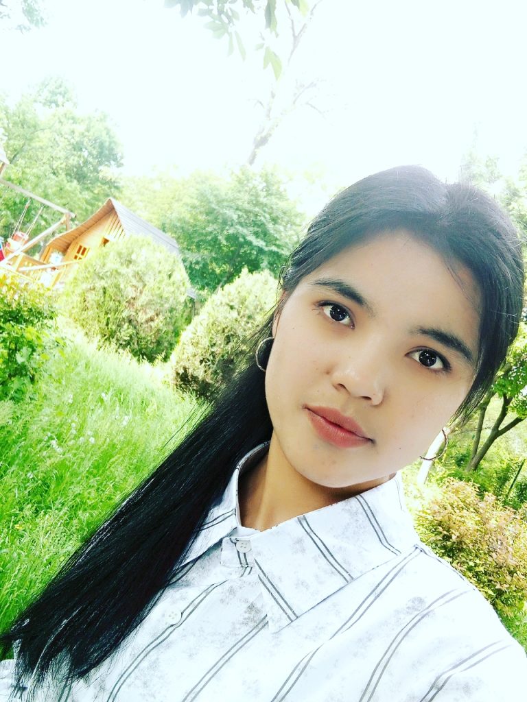 Young Central Asian woman with long dark hair in a ponytail, brown eyes, small hoop earrings, and a white collared shirt with black stripes, posing in front of a lawn with tall grass and trees and bushes and a house and swing set.