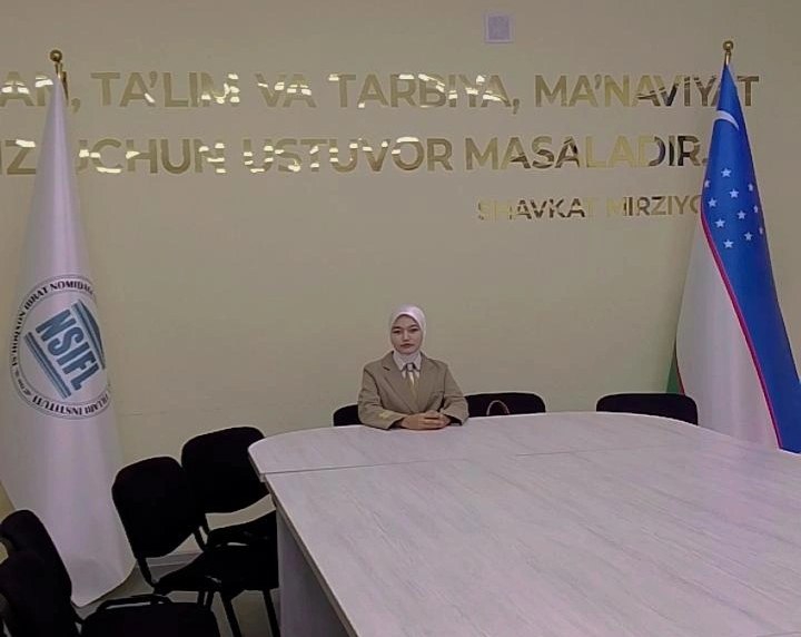 Central Asian woman in a light colored headscarf and tan coat seated at the end of a long table. Flags at either side of her, gold colored Uzbek text on the wall.
