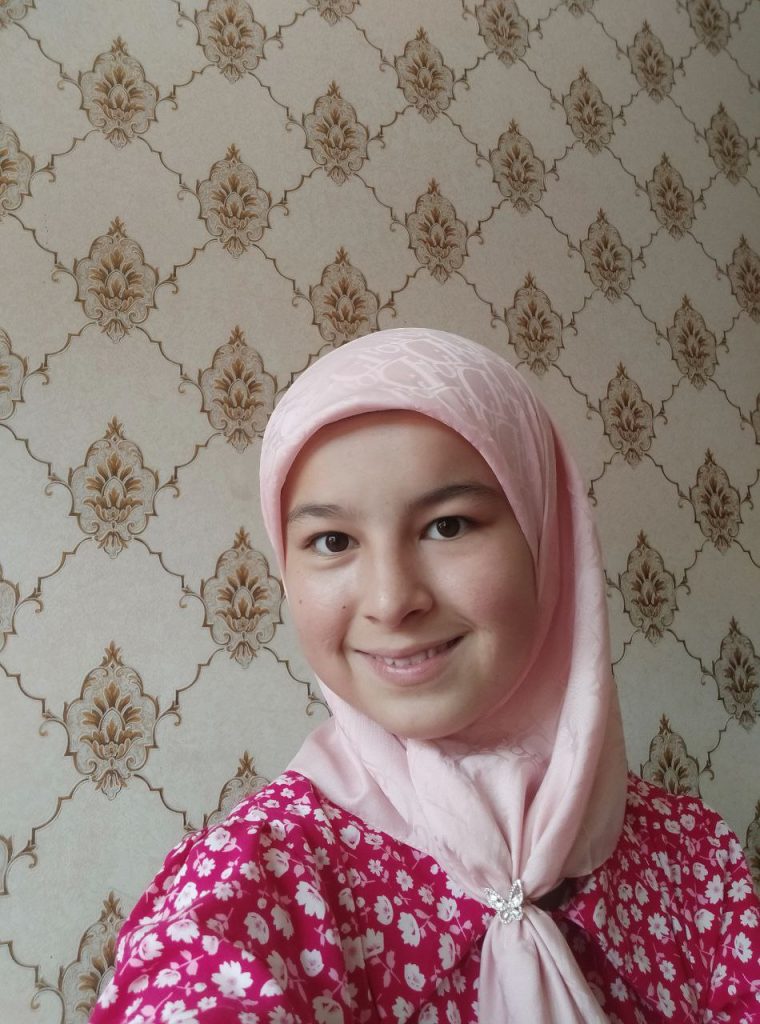 Central Asian teen girl with a pink headscarf and a pink and white floral top poses in front of patterned wallpaper. She's smiling. 