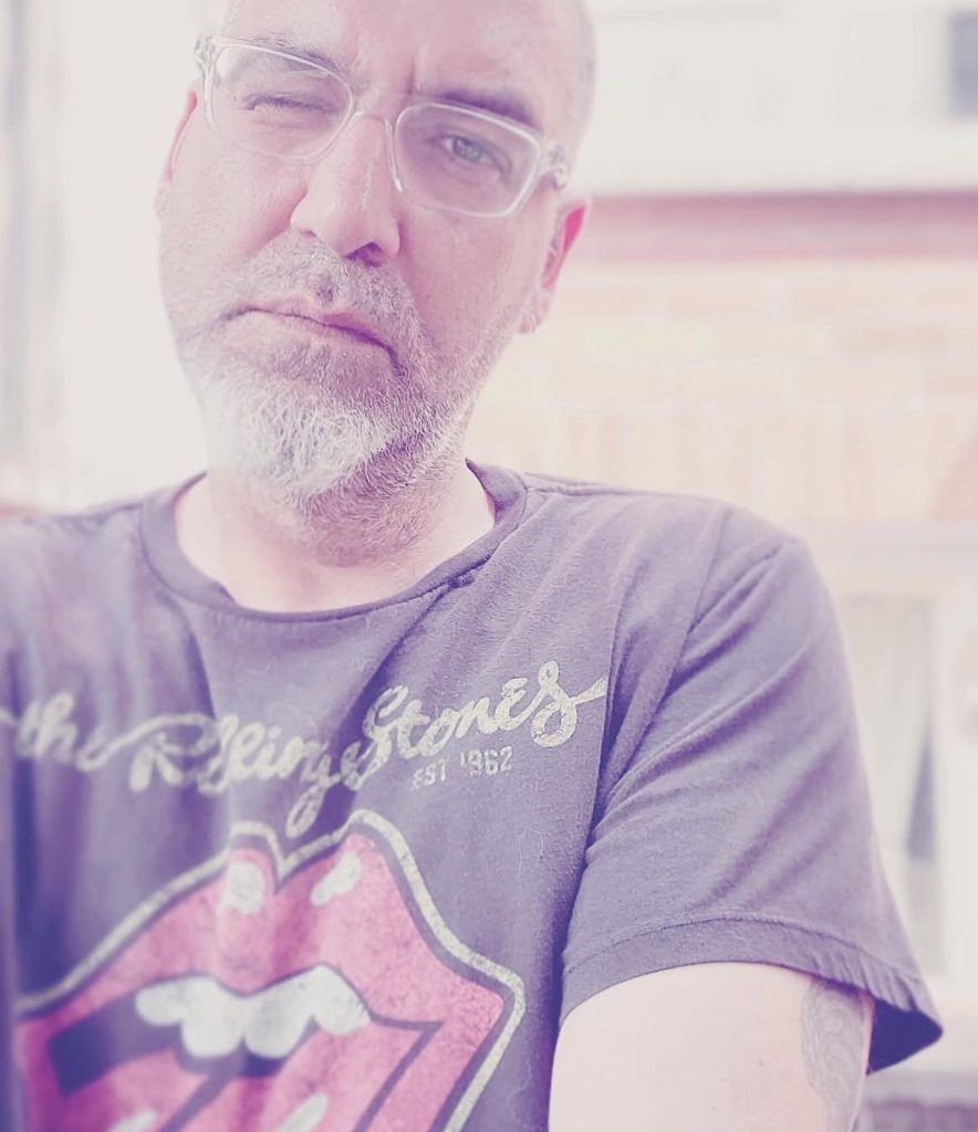 Older middle aged Canadian man winking under his reading glasses. Hazy background, he's wearing a tee shirt. 