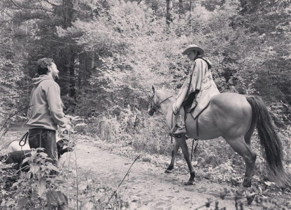 Sepia photograph of a man on horseback in a long blanket and hat riding past some trees talking to another man on food with a dog. 