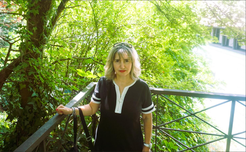 Middle aged European woman with light blonde hair, a black purse, and a black dress with white trim, on a balcony in front of a leafy tree.