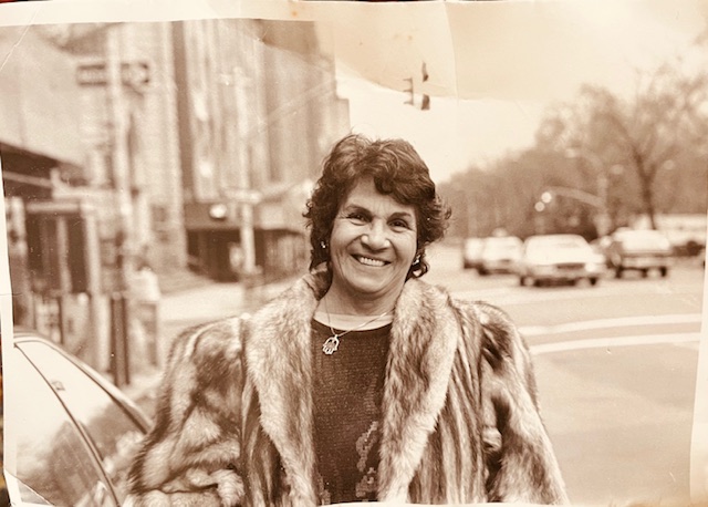 Sepia toned photo of a middle aged woman with curly brown hair, a necklace, and a fur coat over a dark blouse. She's standing next to a car on a city street with buildings and streetlights in the background.