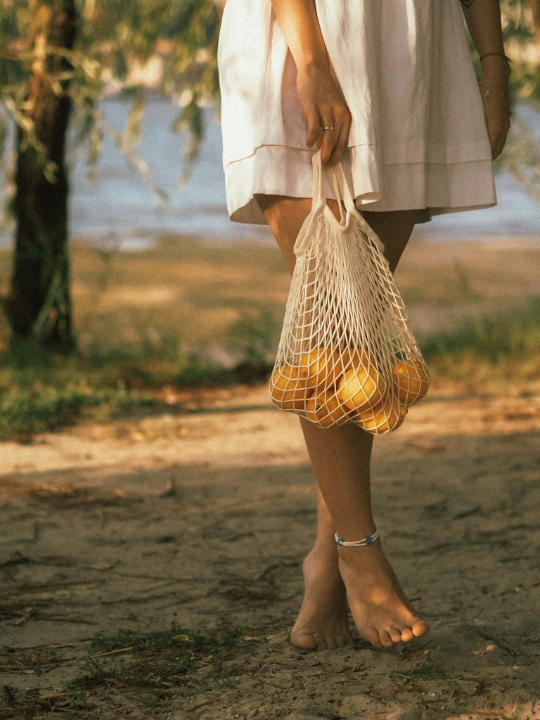 Light skinned barefoot woman stands on grass near trees and water in a light sundress holding a mesh bag of oranges.