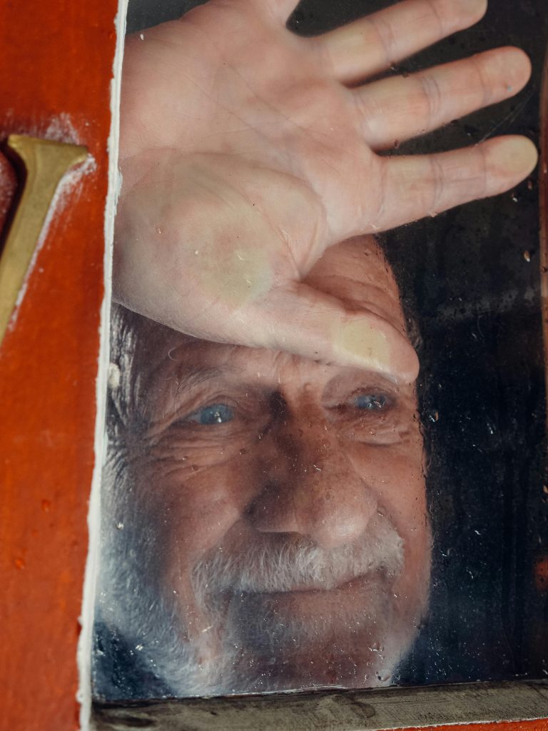 Wistful old light-skinned man with blue eyes stares out a window.