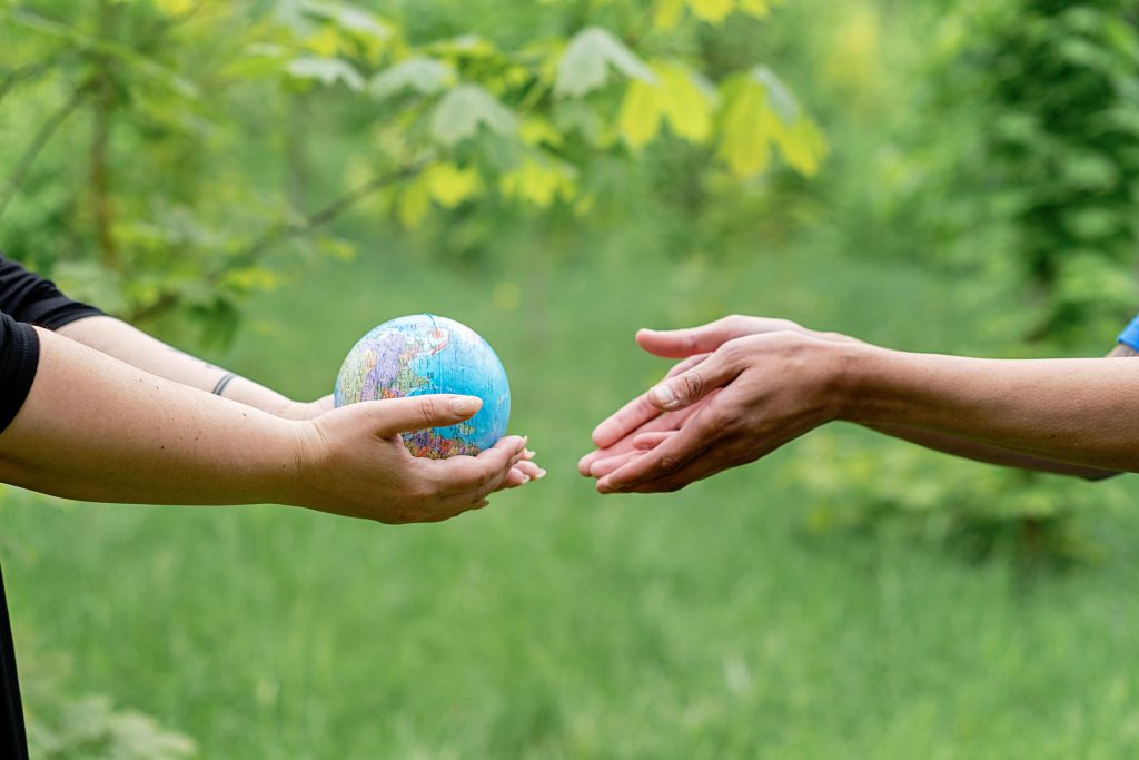 One pair of hands holding a globe to another outstretched pair of hands. Leafy trees and grass in the background.