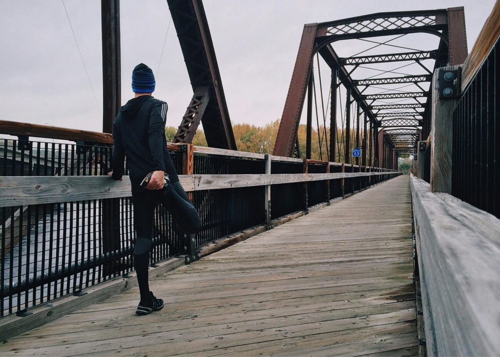 Young man of indeterminate race in a cap, athletic outfit, and shoes stands stretching with his back to us at the beginning of a wooden bridge.