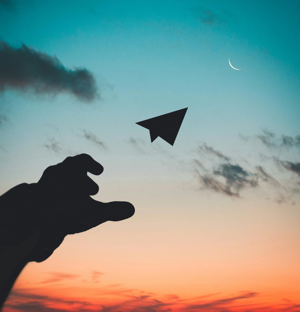 Silhouette of a human hand casting a paper airplane into the sky at sunrise or sunset.