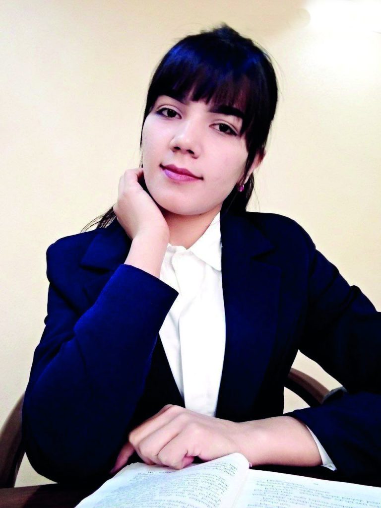 Young Central Asian woman in a dark coat and white blouse seated at a dark wooden table in front of an open book.